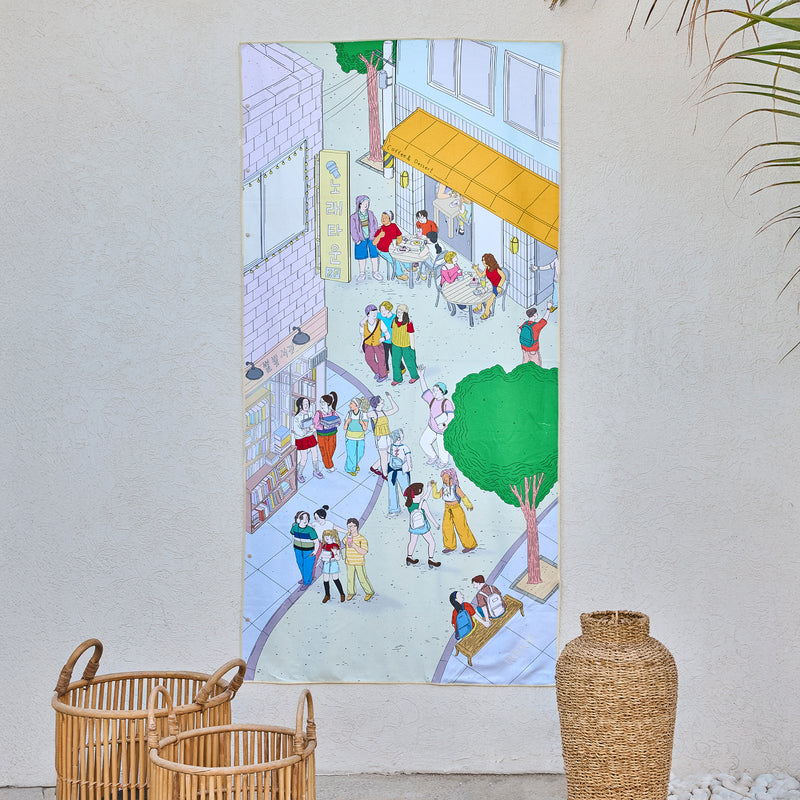 Cherished Memories art beach towel by Joyce Cho, a busy street of cafés and students, hanging on a wall