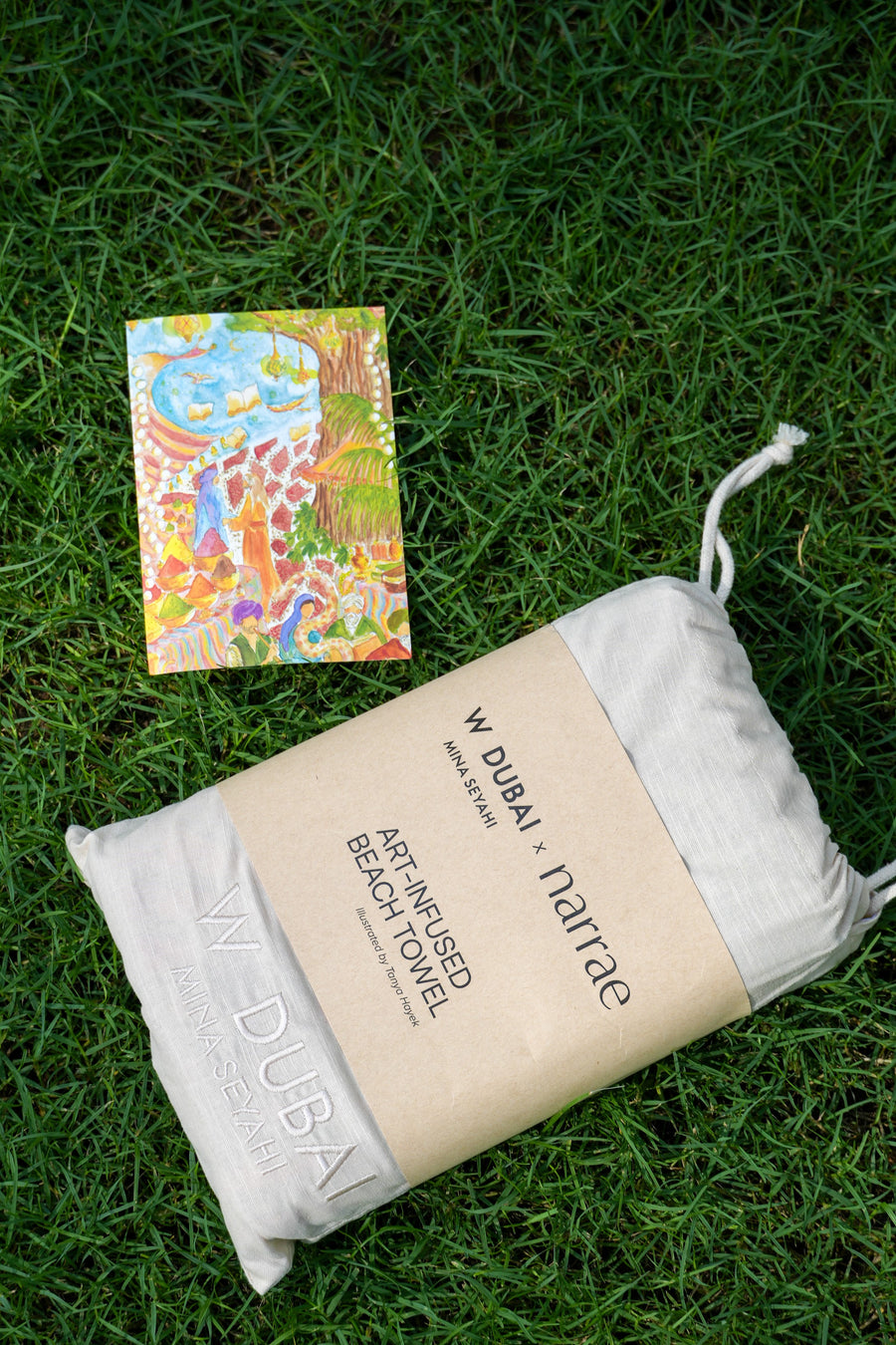 Printed art card on the grass beside the W Dubai x narrae towel in its drawstring bag